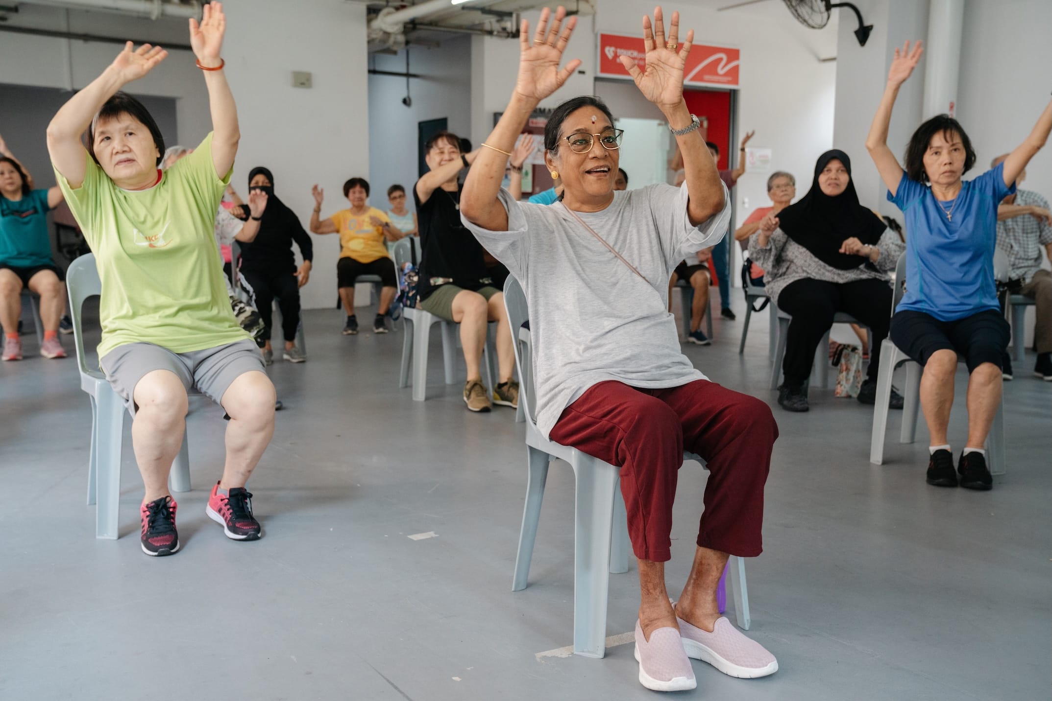 Group of seated adults with arms raised, doing exercise in a light room.